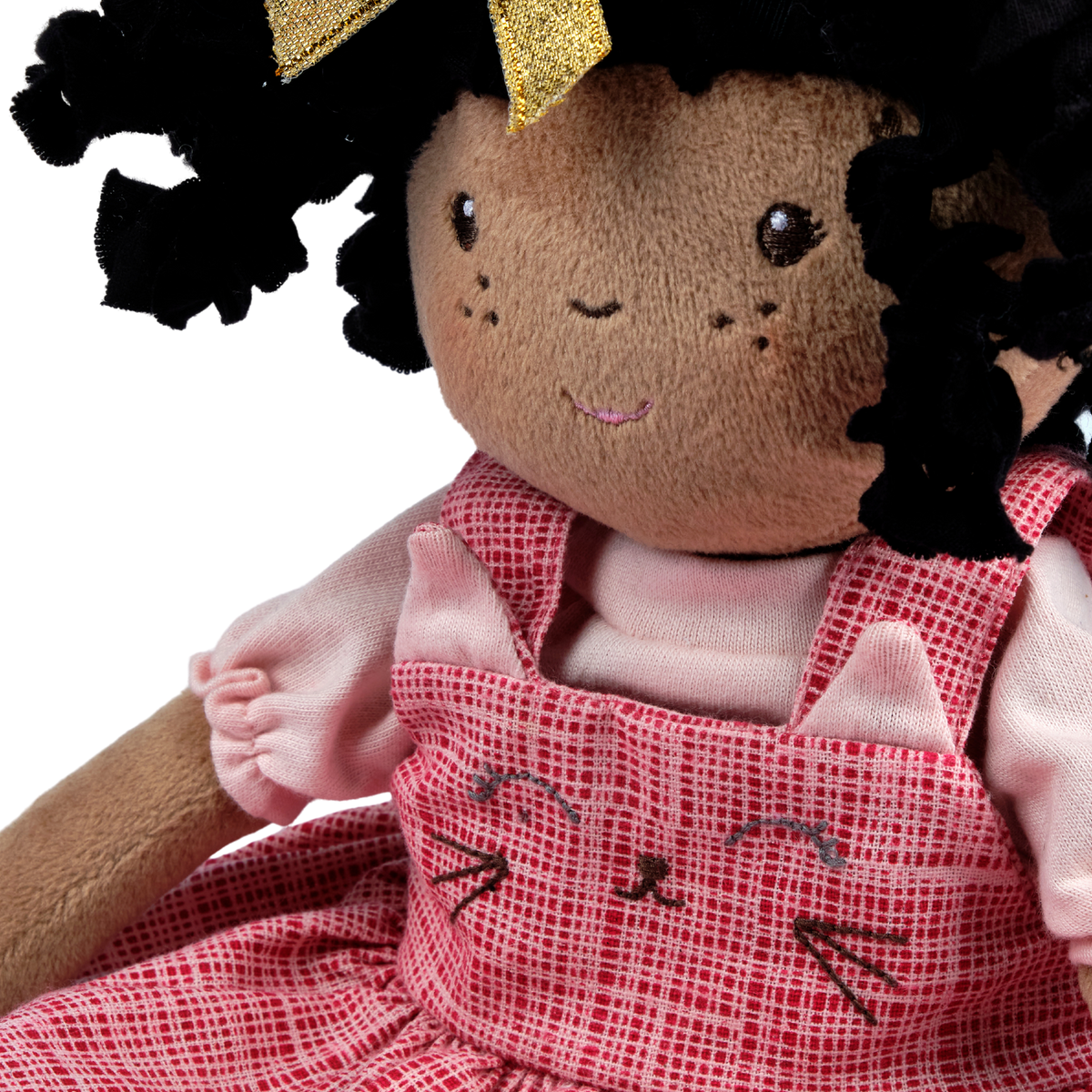 A close-up of a plush doll with dark brown skin, black curly hair with a gold bow, wearing a pink and red checkered dress with embroidered cat faces.