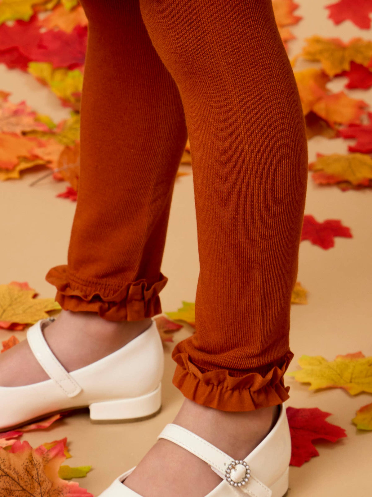 Close-up of a child's legs wearing rust-orange ribbed tights with ruffled cuffs and white Mary Jane shoes, standing on a beige floor scattered with autumn leaves.