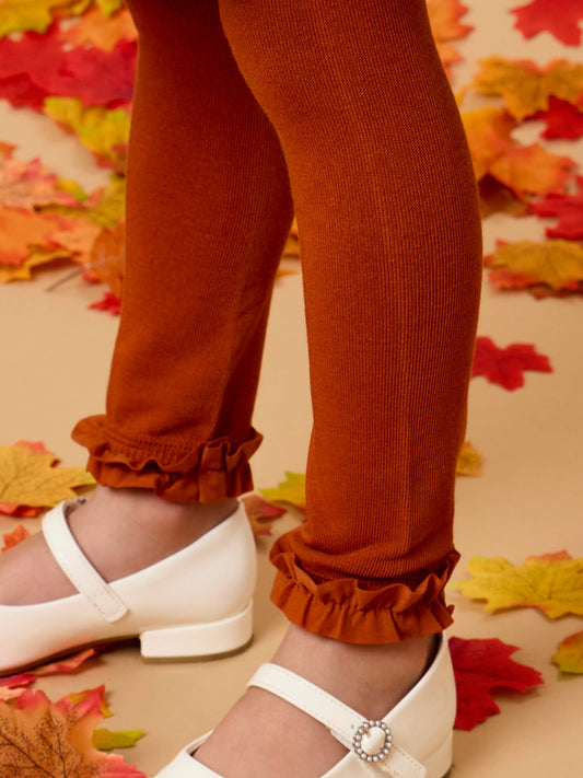 Close-up of a child's legs wearing rust-orange ribbed tights with ruffled cuffs and white Mary Jane shoes, standing on a beige floor scattered with autumn leaves.