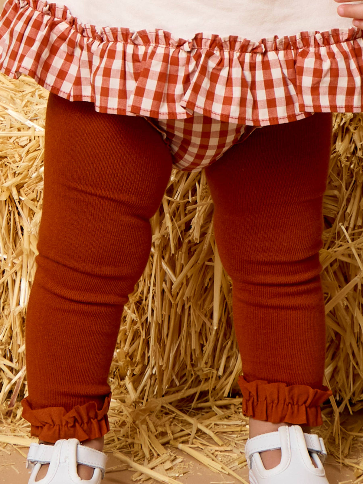 A close-up of a child's lower body wearing rust-colored ribbed leggings with ruffled cuffs, a red-and-white gingham ruffled skirt, and white shoes, standing in front of a hay bale backdrop.