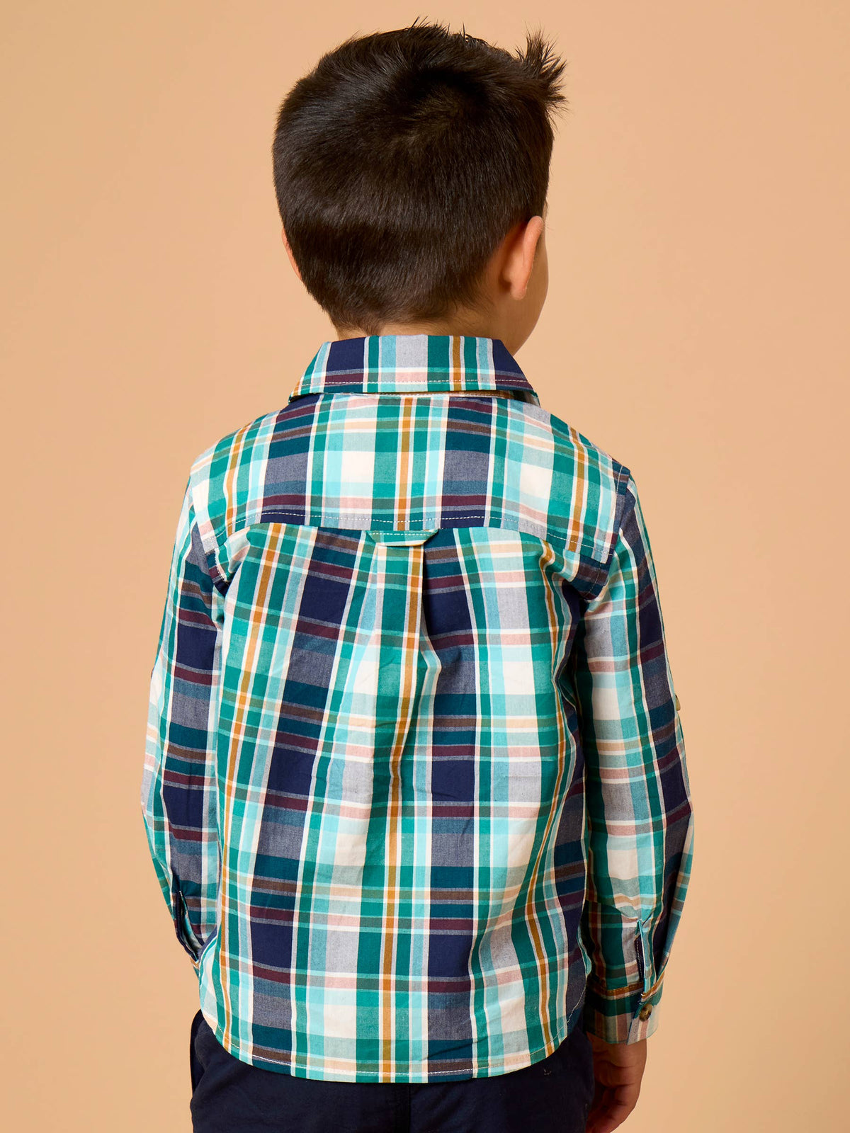 A child viewed from behind wearing a long-sleeve plaid button-up shirt in navy, teal, green, white, and thin orange stripes, set against a solid peach background.