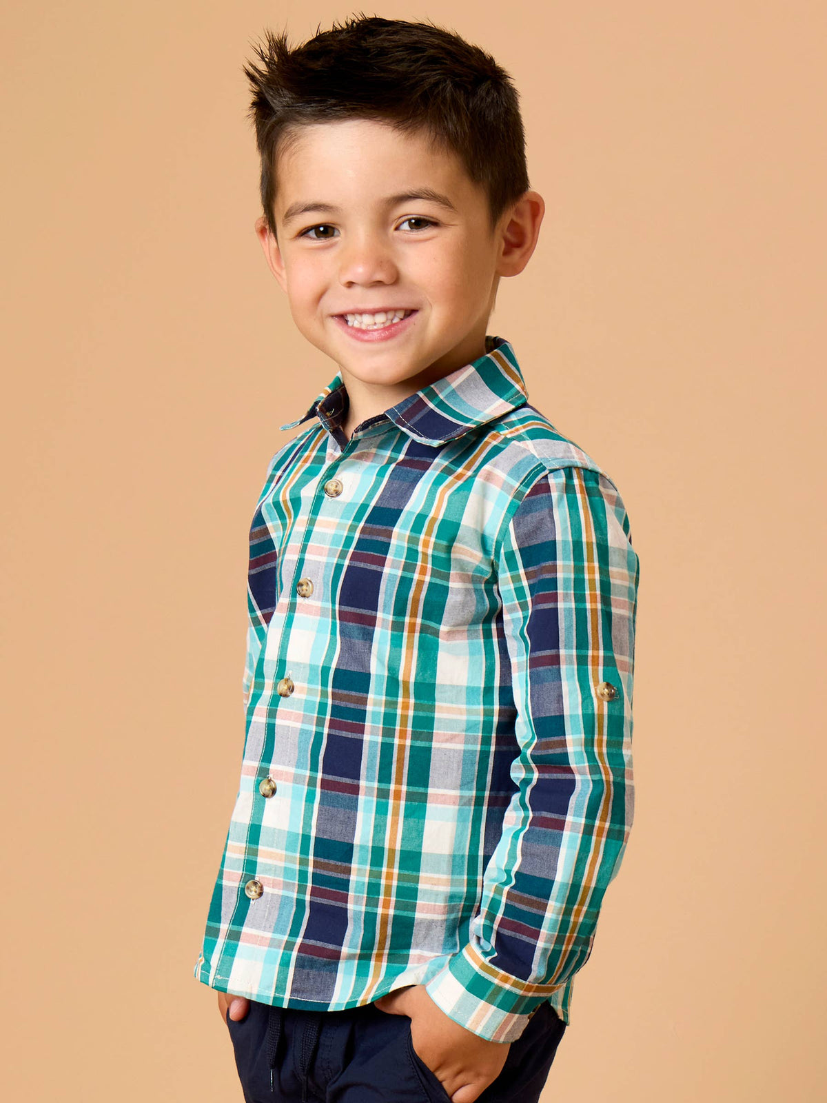 A young boy smiling and posing in a long-sleeve plaid button-down shirt with green, navy, white, and orange stripes, set against a solid beige background.
