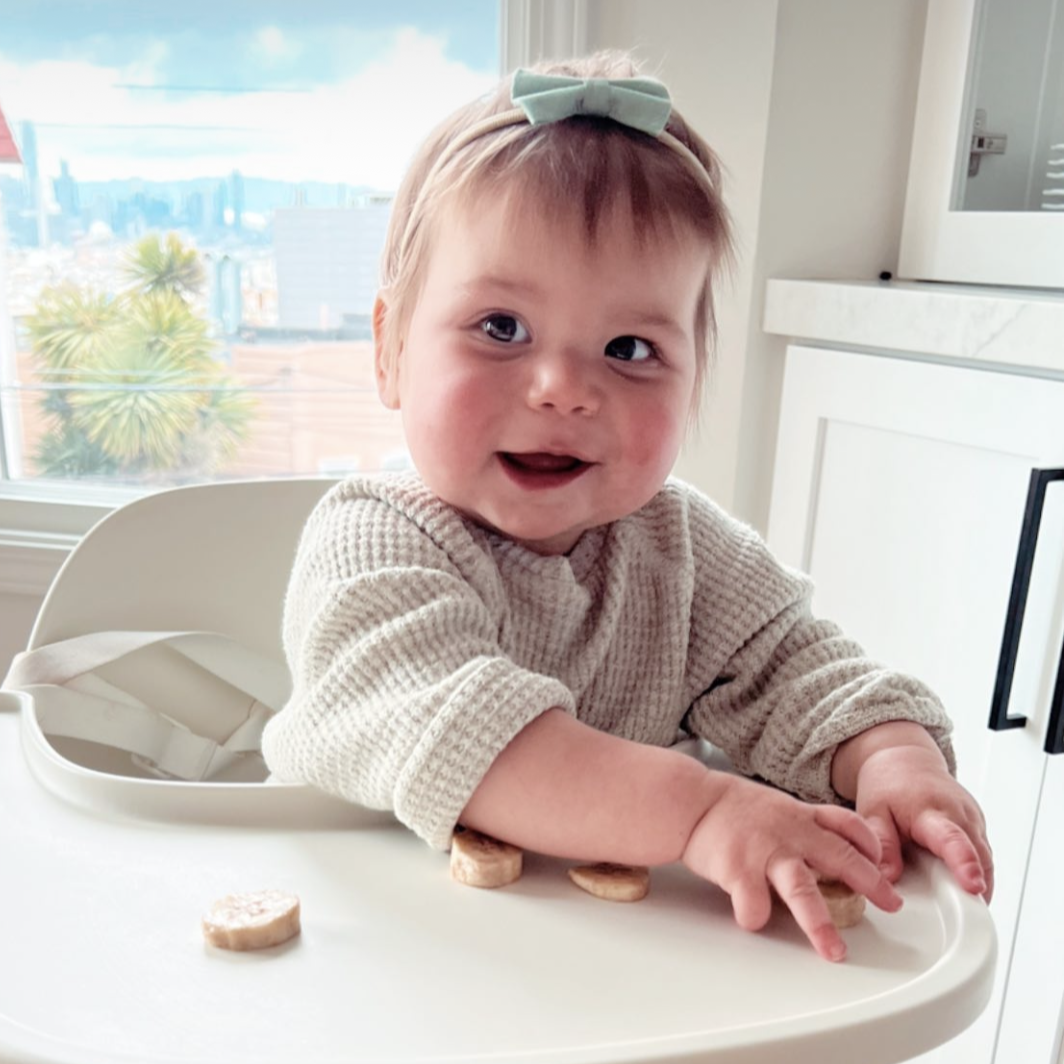 Baby sitting in a high chair with a bow headband, smiling in a bright kitchen.