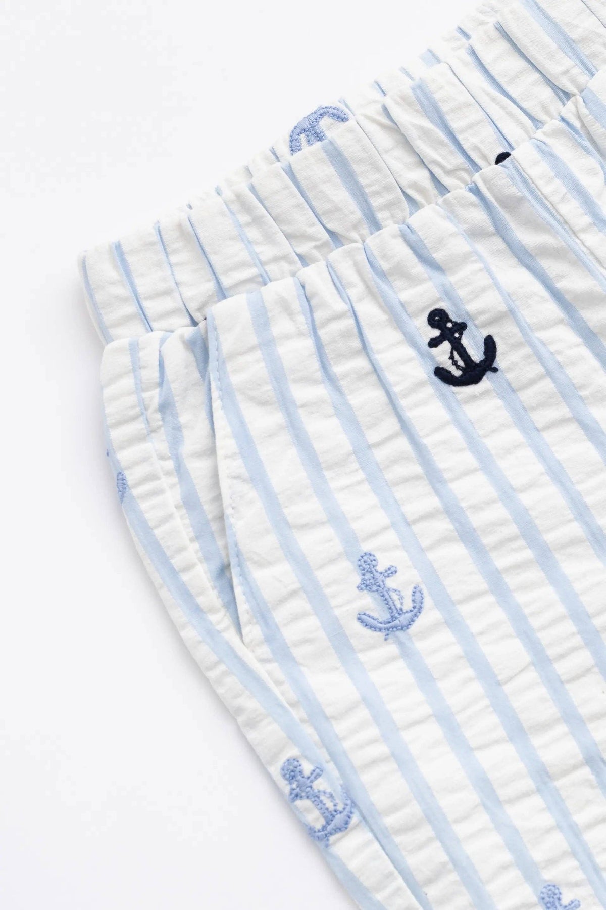 Close-up of a pair of white and light blue vertically striped cotton pajama shorts with embroidered anchor motifs in navy and light blue.