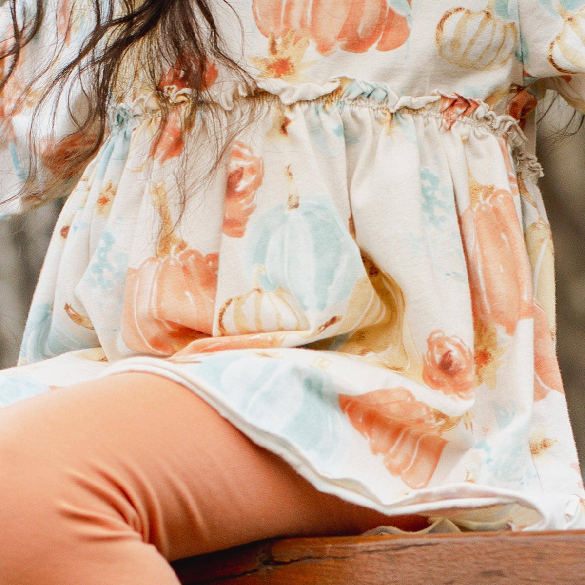 A close-up of a child wearing a cream-colored dress with a watercolor pumpkin print in peach, orange, and light blue, paired with peach leggings, sitting on a wooden bench.