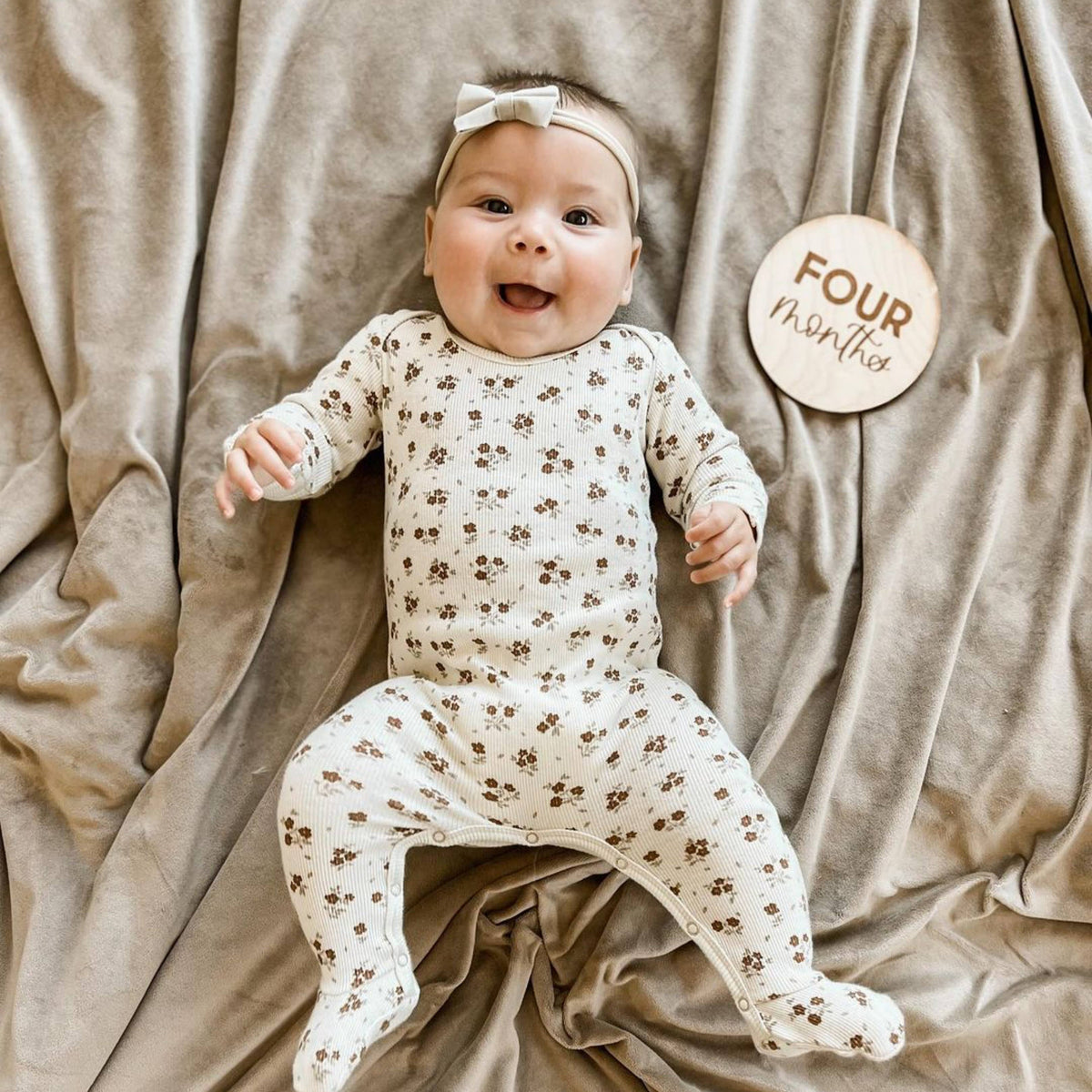 Baby in a floral onesie sitting on a beige blanket with a 'Four Months' milestone card.