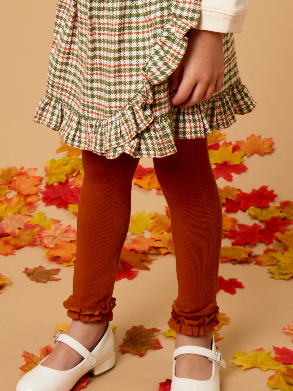 A child wearing a green, red, and white plaid ruffled skirt, rust-brown tights, and white Mary Jane shoes, standing on a beige floor scattered with autumn leaves.