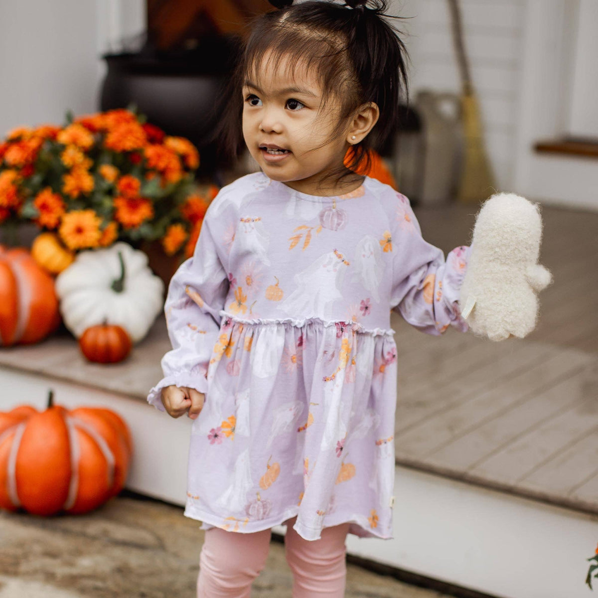 Young girl in a light purple dress with floral patterns holding a white fluffy toy, surrounded by pumpkins and flowers.