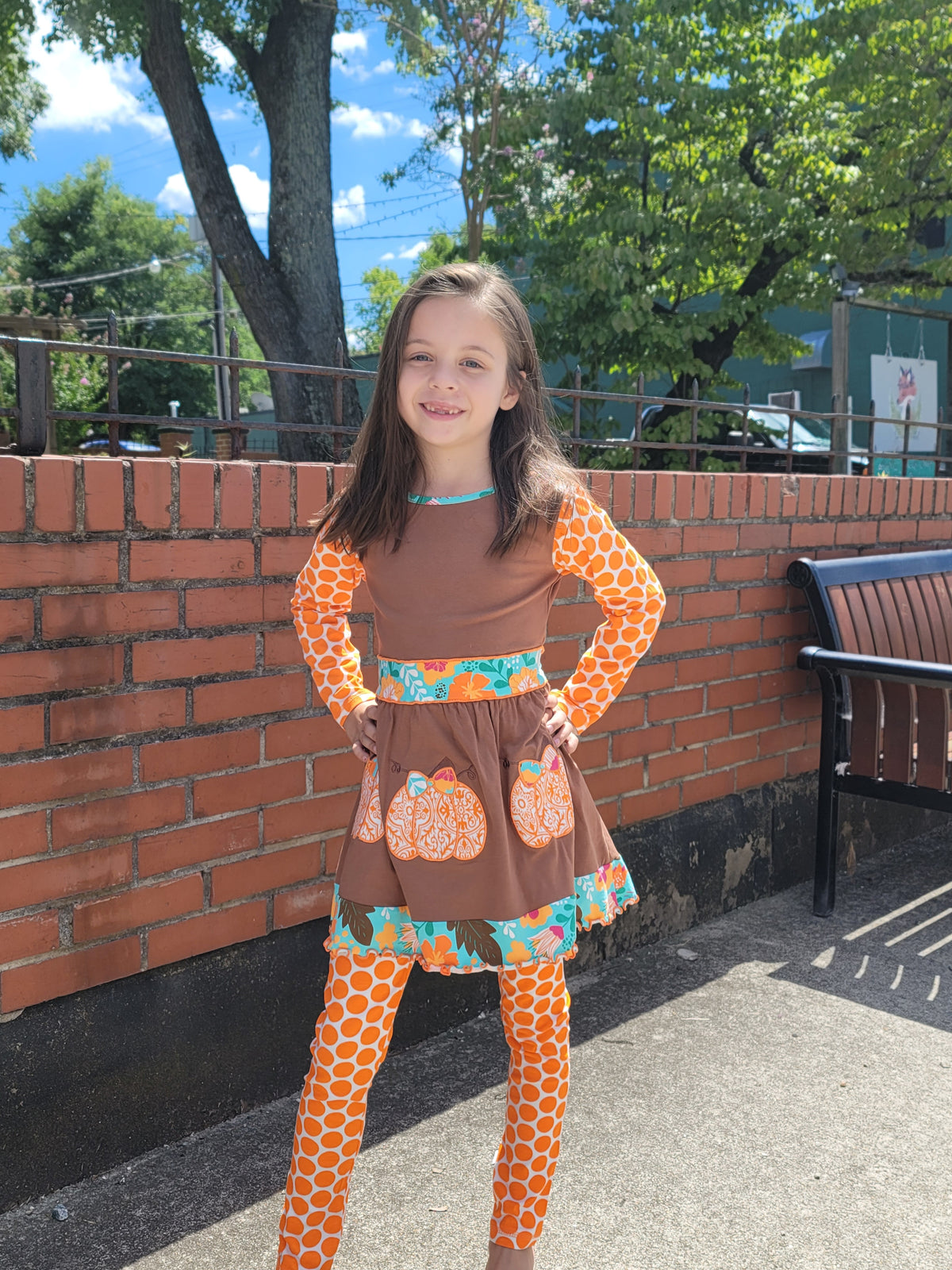 Child wearing a brown dress with orange pumpkin patterns on a sidewalk.