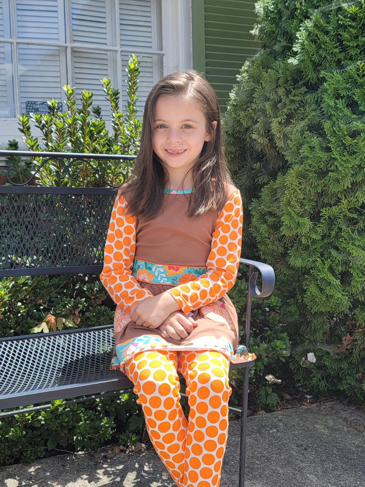 Child seated outdoors wearing Ann Loren’s brown pumpkin tunic with orange polka-dot sleeves and matching leggings.