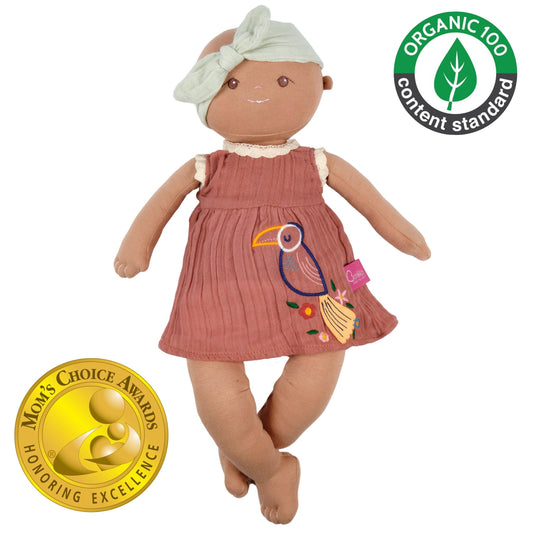 A doll wearing a pink dress with embroidered toucan is against a white background with a gold mom's choice award logo and a green and grey organic logo in the other corner. 