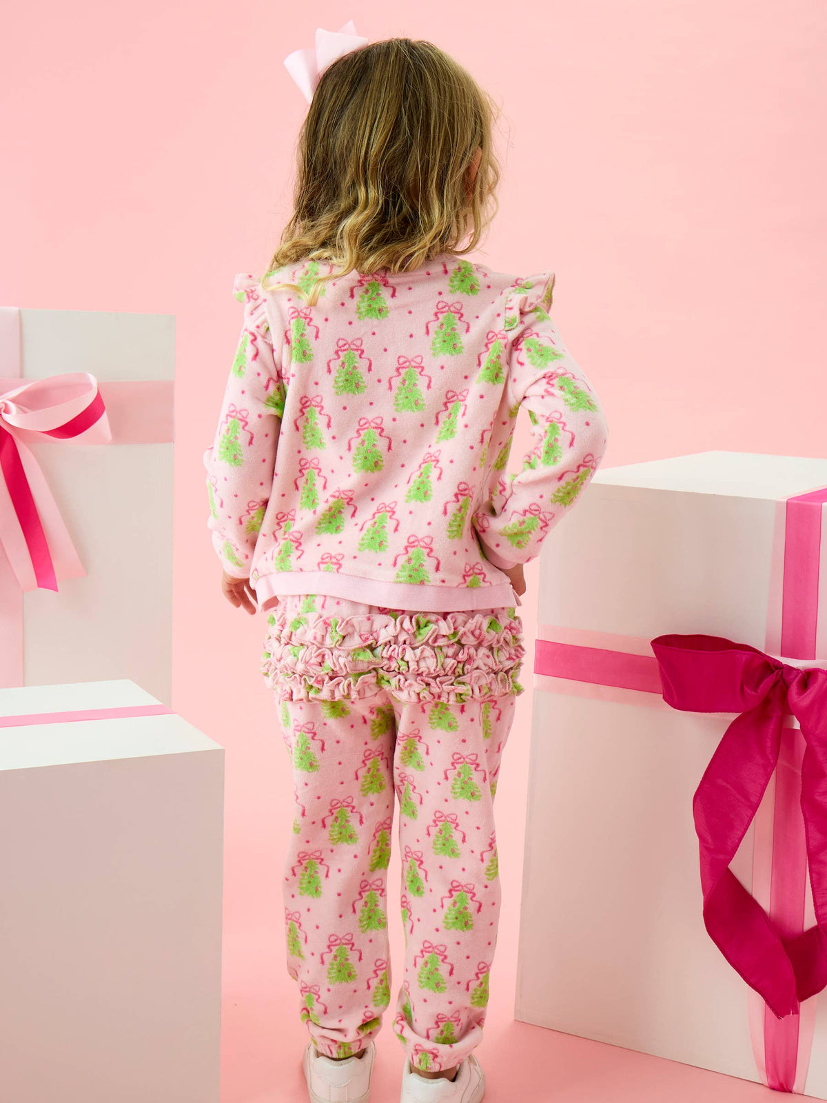A young girl viewed from behind wearing a pink pajama set with a green and white Christmas tree pattern, standing between large white gift boxes with pink ribbons against a pink background.