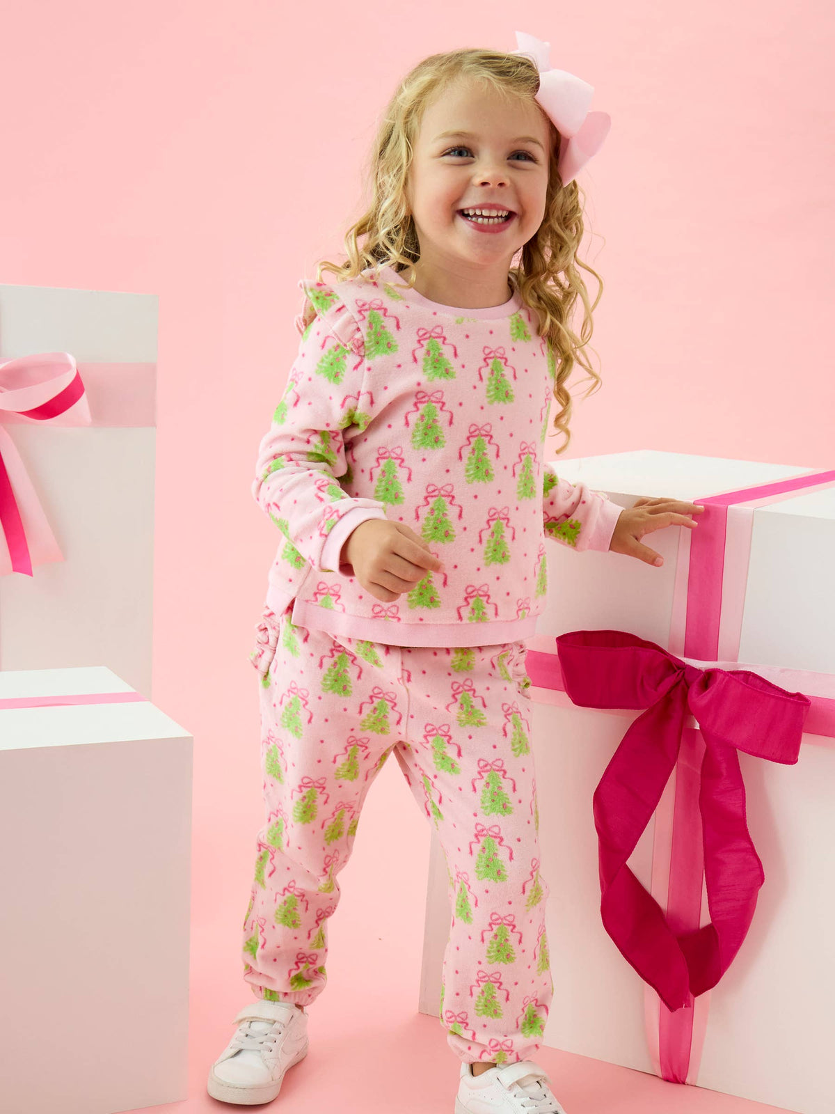 Girl in a pink sweatshirt & sweatpants set with green christmas tree pattern surrounded by christmas gifts