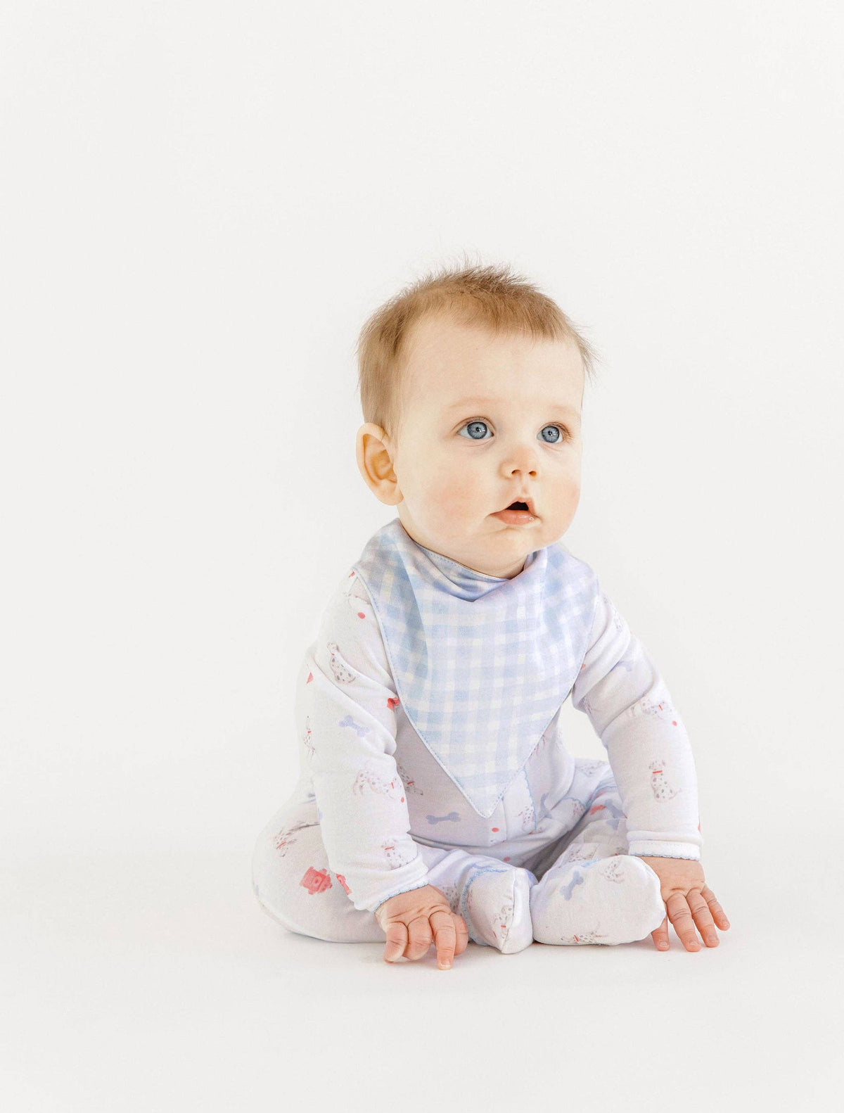 Baby sits against a white background wearing a onesie with a dalmation dog print and a blue checked bandana. 