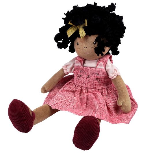 A plush doll with black curly hair, a gold bow, a pink plaid dress over a light pink shirt, and dark red shoes, shown against a plain white background.