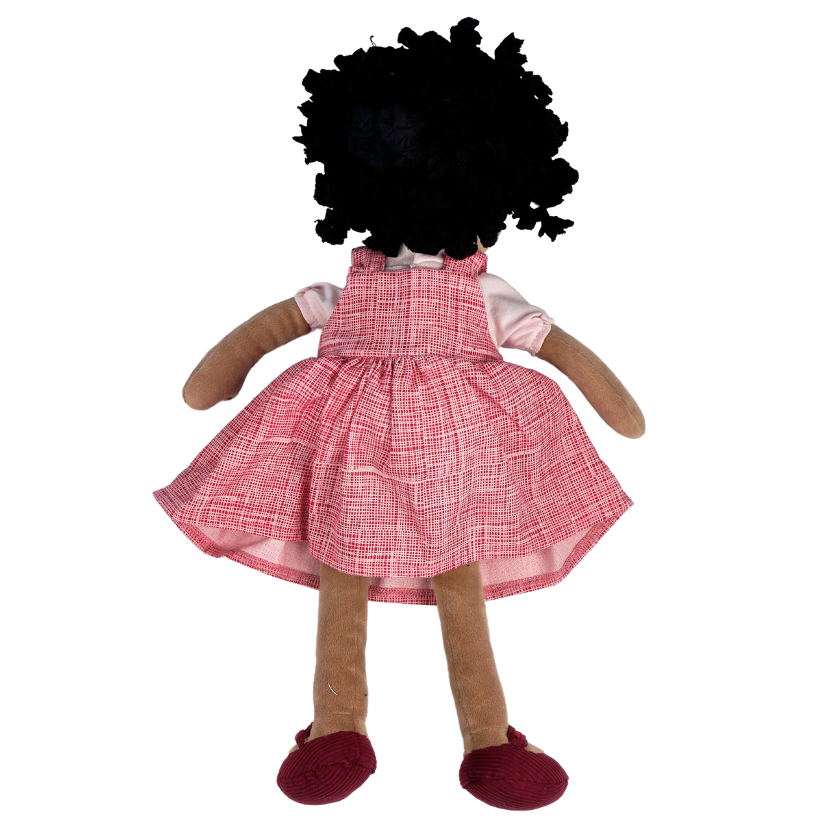 A plush doll viewed from the back, featuring a black curly wig, tan fabric skin, a pink and white plaid pinafore dress, a light pink shirt, and dark red shoes, isolated on a white background.