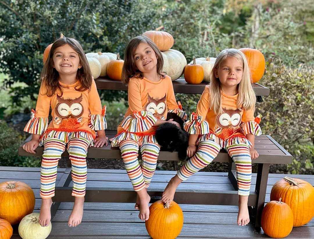 Three children in matching outfits with owl designs sitting on a wooden bench surrounded by pumpkins.
