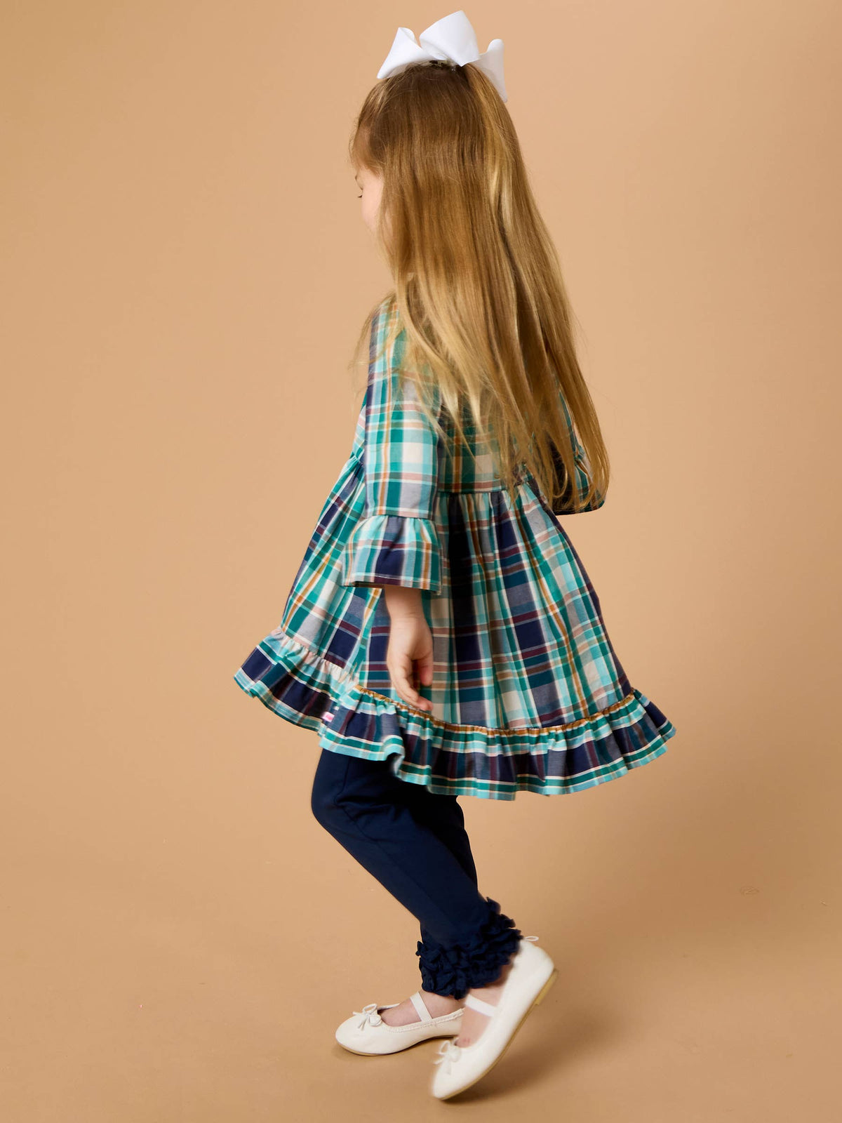 A young girl with long blonde hair wearing a green and navy plaid ruffled dress, navy leggings, and white shoes, shown from a side-back angle against a solid tan studio background.