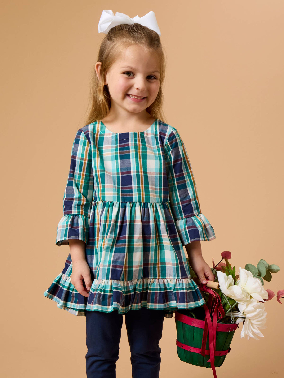 A young girl with a white bow in her hair stands against a solid beige background, wearing a green, blue, and white plaid long-sleeve dress with ruffled cuffs and hem, paired with navy blue pants, and holding a small green and red flower basket.