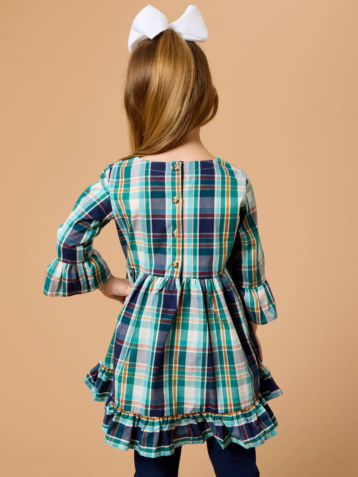 A young girl viewed from behind wearing a plaid long-sleeve dress with a ruffled hem and back buttons, paired with a large white bow in her hair, against a solid tan background.