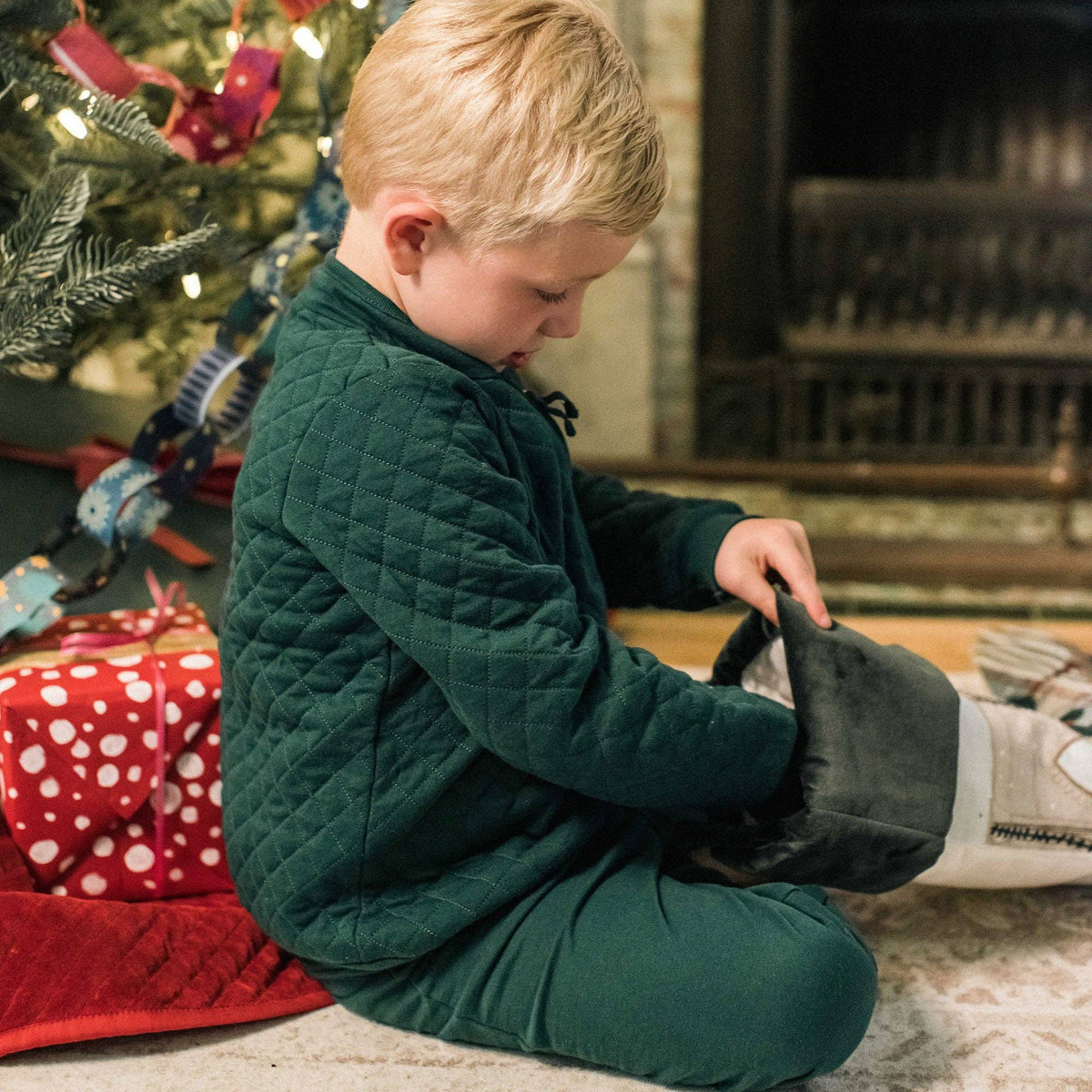 Child sitting under a Christmas tree with a stocking wearing a green quarter zip jogger set that is quilted in texture. 