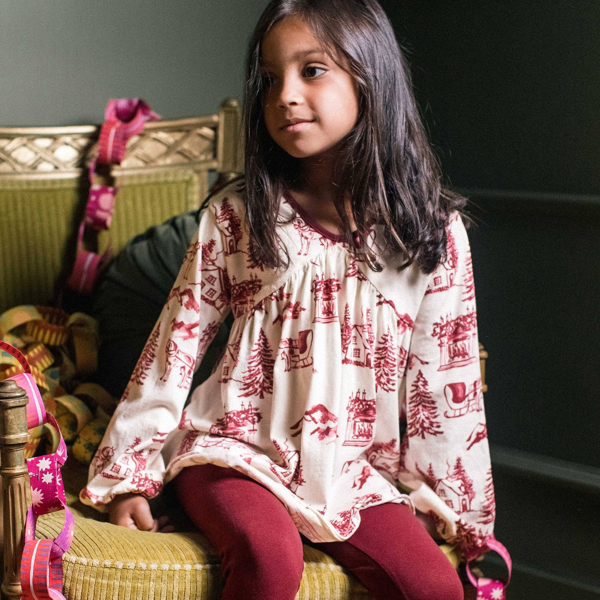 Girl sits on a chair wearing a vintage christmas set with a classic motif fabric for the tunic featuring a winter chalet scene and carriages pulled by horses, paired with a wine colored legging. 