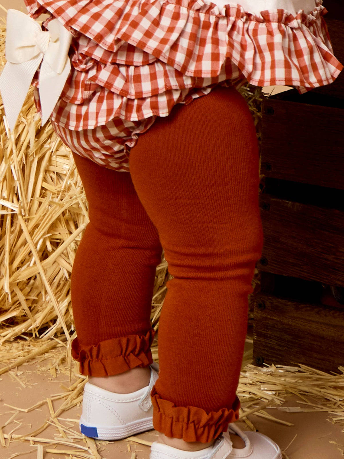 Toddler's lower body wearing rust-orange ribbed knit leggings with ruffled cuffs, a red-and-white gingham ruffled top, and white sneakers, standing on a floor with straw and a wooden crate in the background.