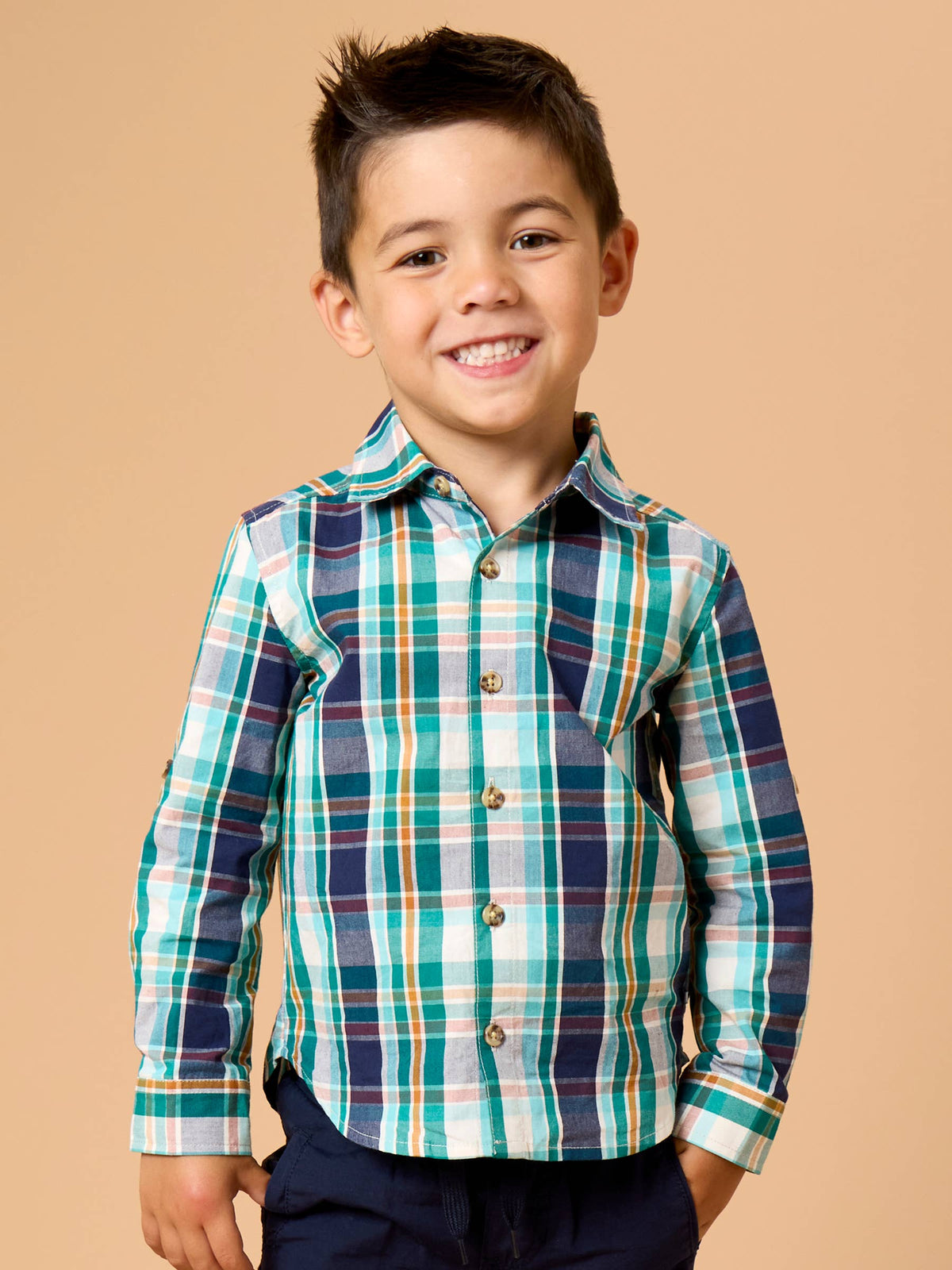 A young boy smiling and wearing a long-sleeve plaid button-up shirt in shades of teal, navy, green, and white, paired with dark pants, set against a solid beige background.