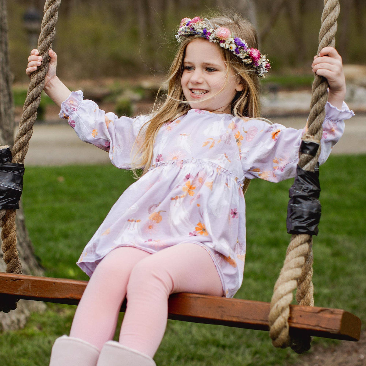 Young girl in a floral dress and flower crown sitting on a swing outdoors.