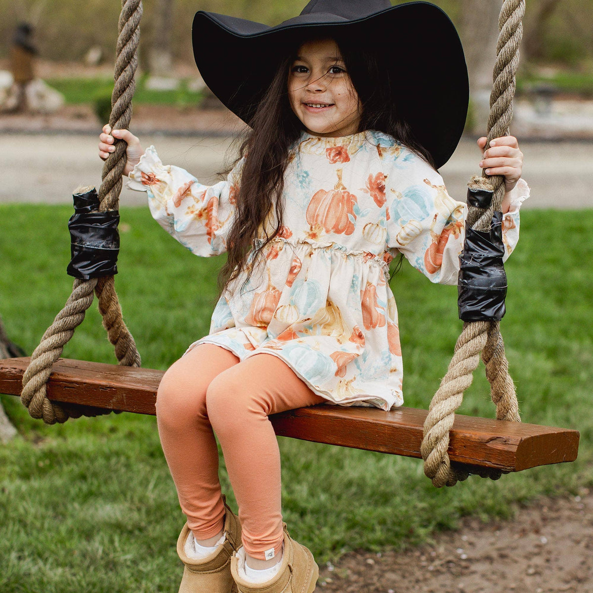 Child sitting on a swing wearing a floral dress and wide-brimmed hat in an outdoor setting.