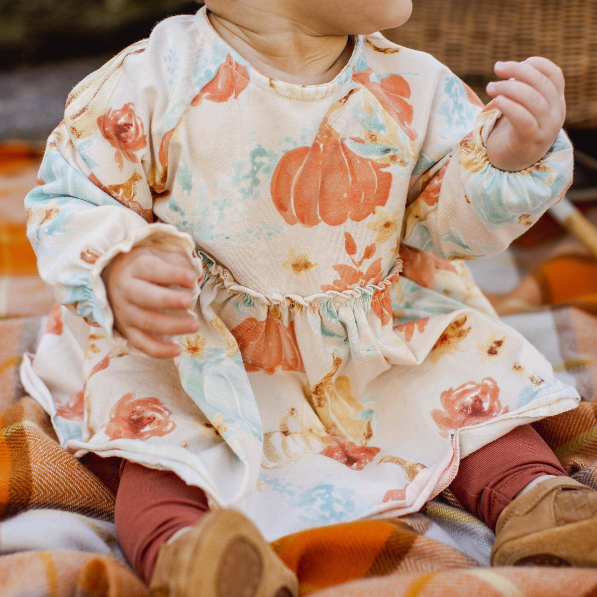 A baby wearing a long-sleeve cream dress with a watercolor-style orange pumpkin and floral print, paired with rust leggings and tan boots, sitting on a plaid blanket.