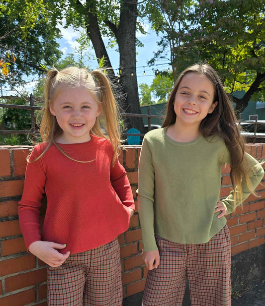 Two young girls standing next to a brick wall with trees and buildings in the background.